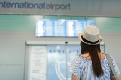 Back rear view of young traveler tourist woman in hat looking on schedule, timetable waiting in lobby hall at international airport. Passenger traveling abroad on weekends getaway. Air flight concept